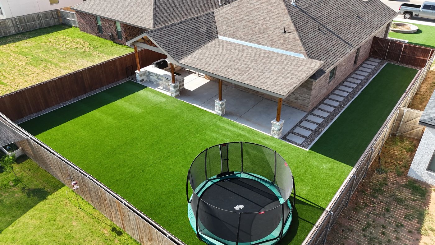 Aerial backyard turf lawn with in-ground trampoline and covered patio outdoor kitchen in Desert Ridge, Odessa TX
